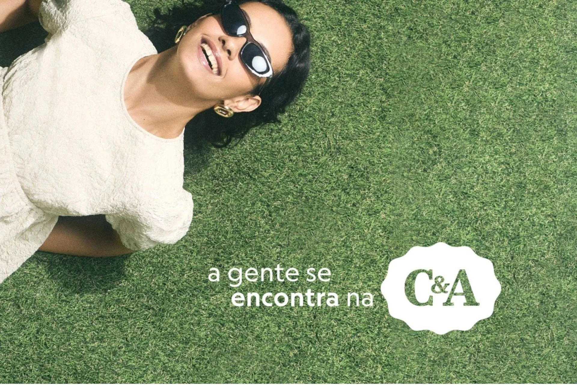 Mulher deitada na grama com óculos escuro e sorriso no rosto, com um vestido de renda. Lê-se o texto 'A gente se encontra na C i A'