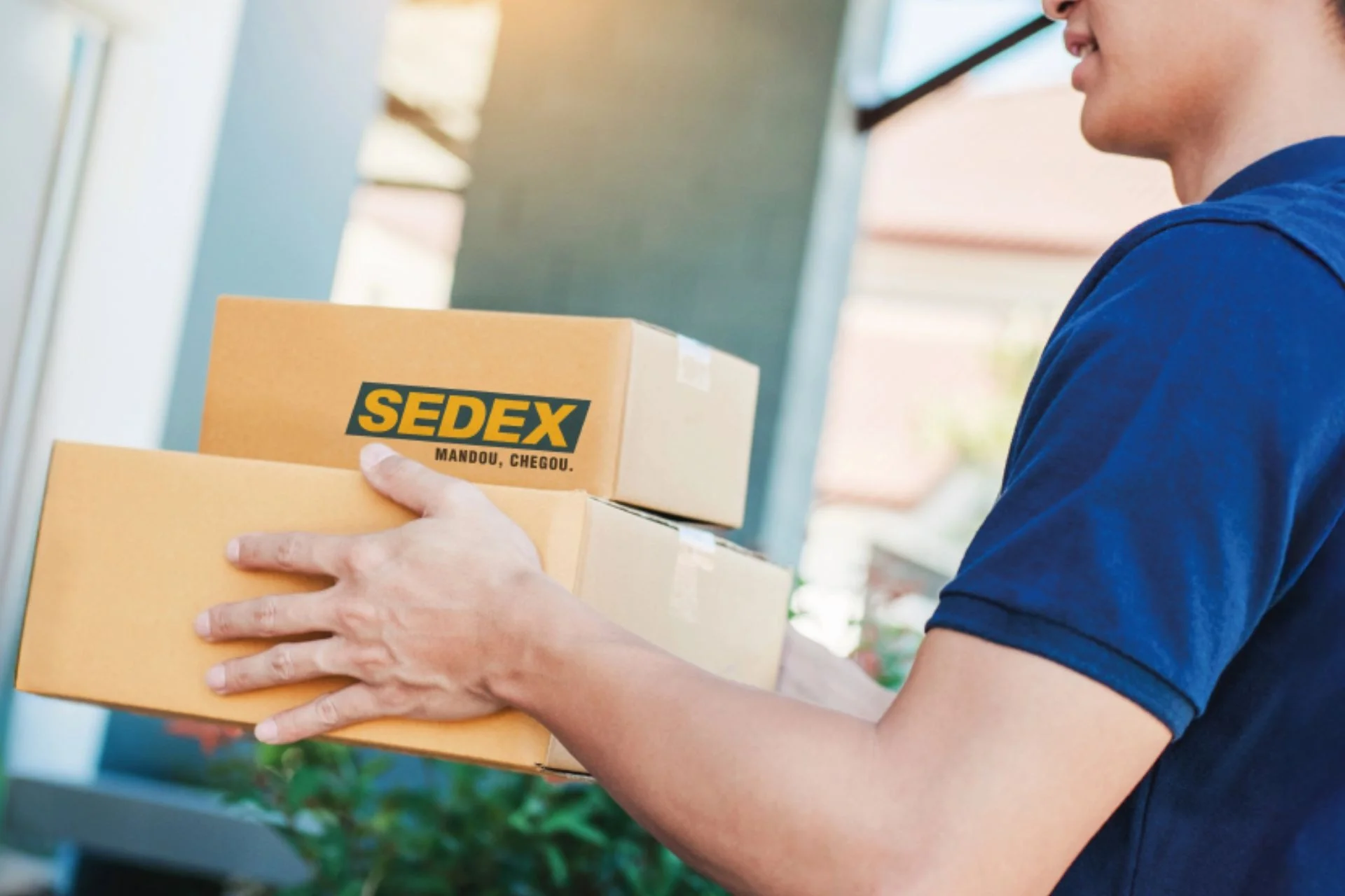 Homem com camisa azul segura pacotes com a marca SEDEX dos Correios.