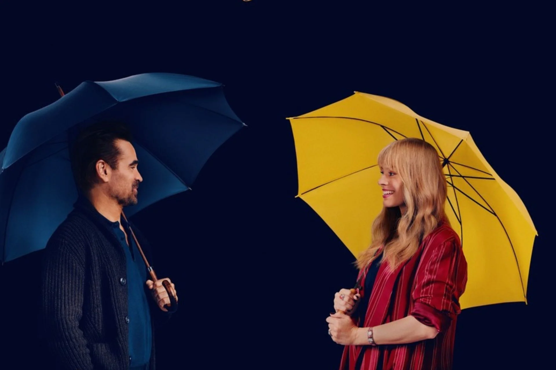 Homem e mulher se encaram sorrindo, o homem está sob um guarda-chuva escuro e a mulher sob um amarelo.