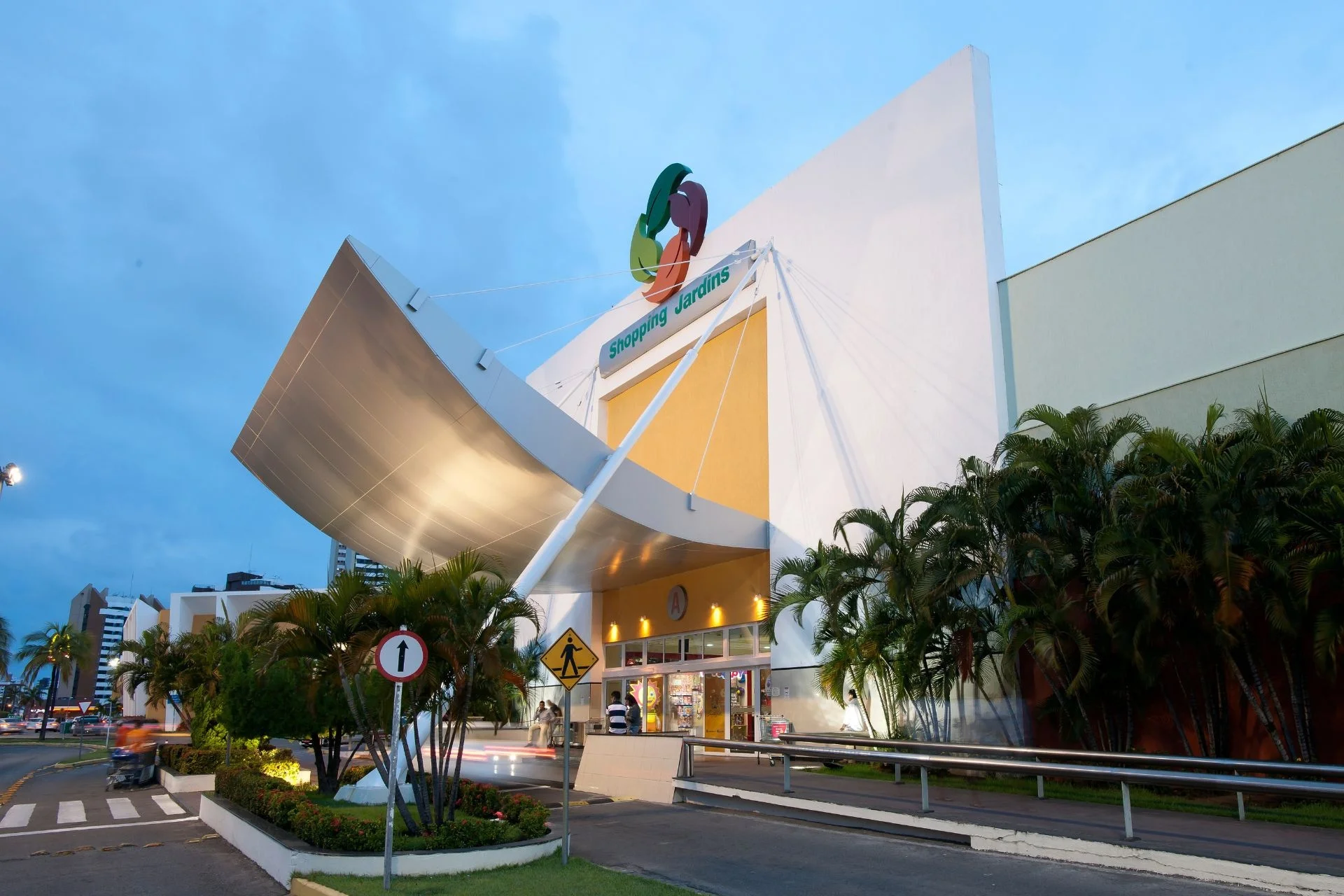 Fachada do Shopping Jardins ao anoitecer, com árvores e pista que leva a entrada A. Vemos prédios ao fundo e um céu azul com algumas nuvens.