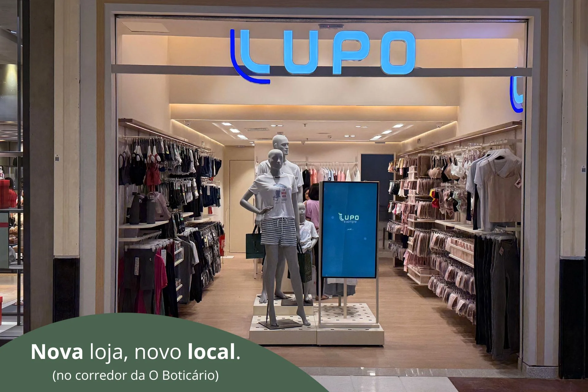 Loja da Lupo vista de frente em um shopping, com letreiro azul "LUPO" na entrada, araras de roupas e lingerie nas laterais e manequins ao centro; no rodapé, um banner verde com o texto "Nova loja, novo local. (no corredor da O Boticário)".